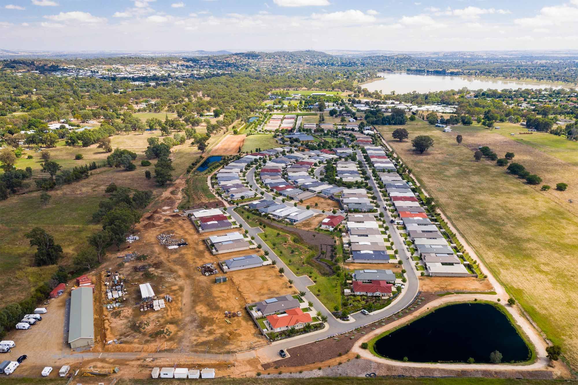 Aerial view of a residential neighborhood under construction, with houses, a small pond, and nearby open fields, with a lake in the background.