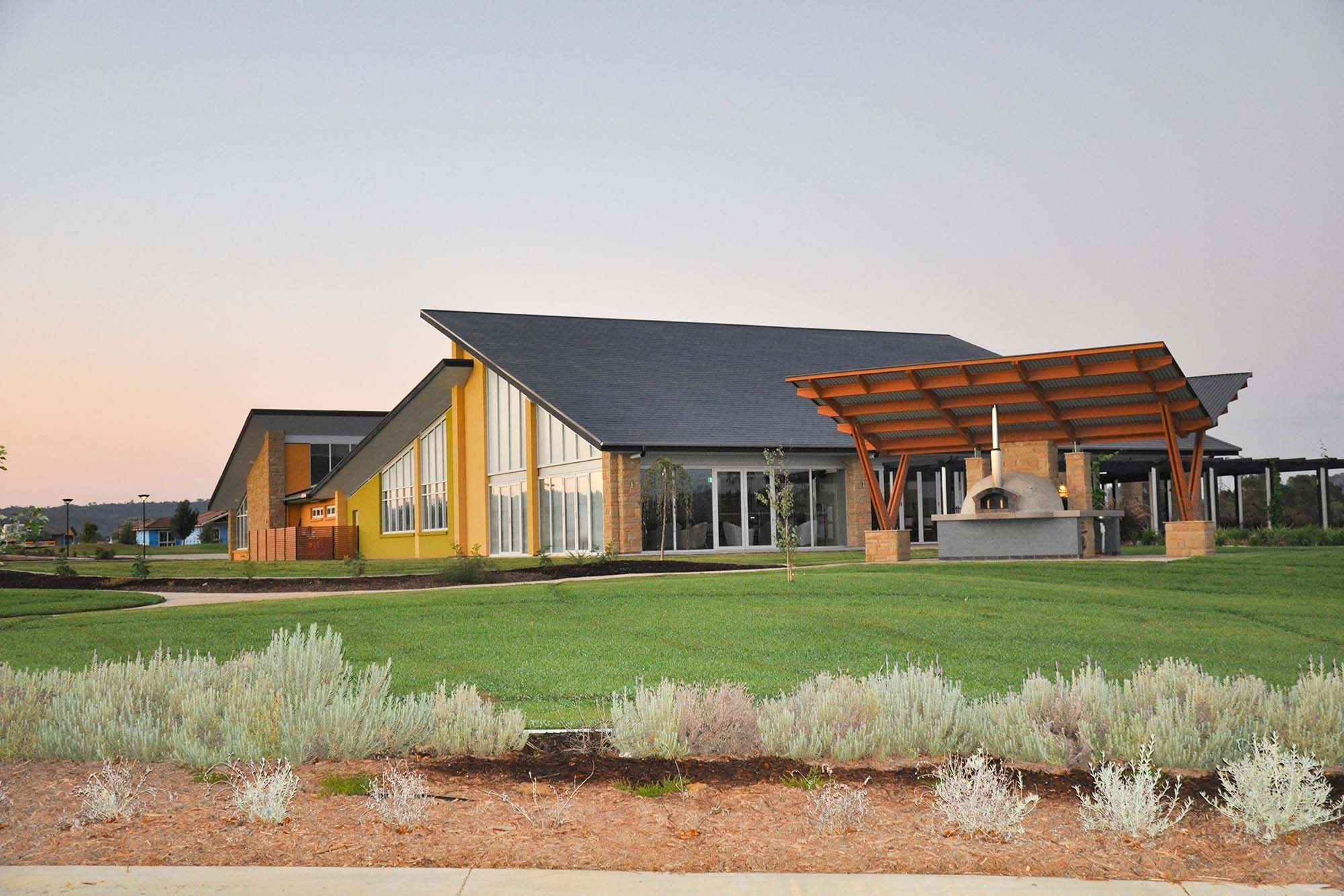 Modern building with large windows and a sloped roof, featuring a covered outdoor kitchen area with a pizza oven, set in a landscaped lawn with bushes and trees.