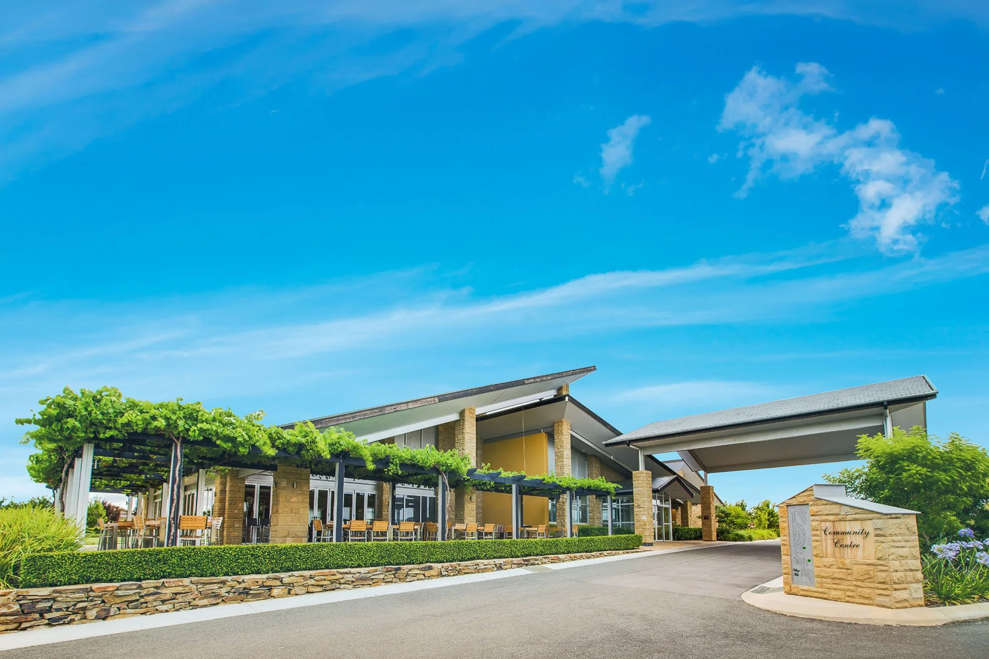 Community center building with outdoor seating area, green vine-covered pergola, stone wall, and a sign reading 'Community Center' against a bright blue sky.