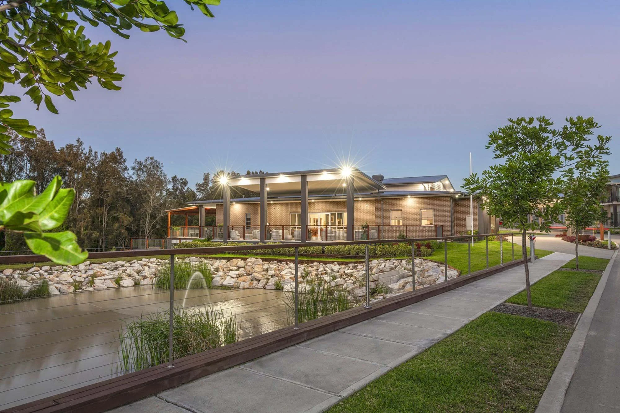 A modern building with exterior lighting, surrounded by trees, a pond with rocks and a fountain, a sidewalk, and manicured grass, during dusk or early evening.
