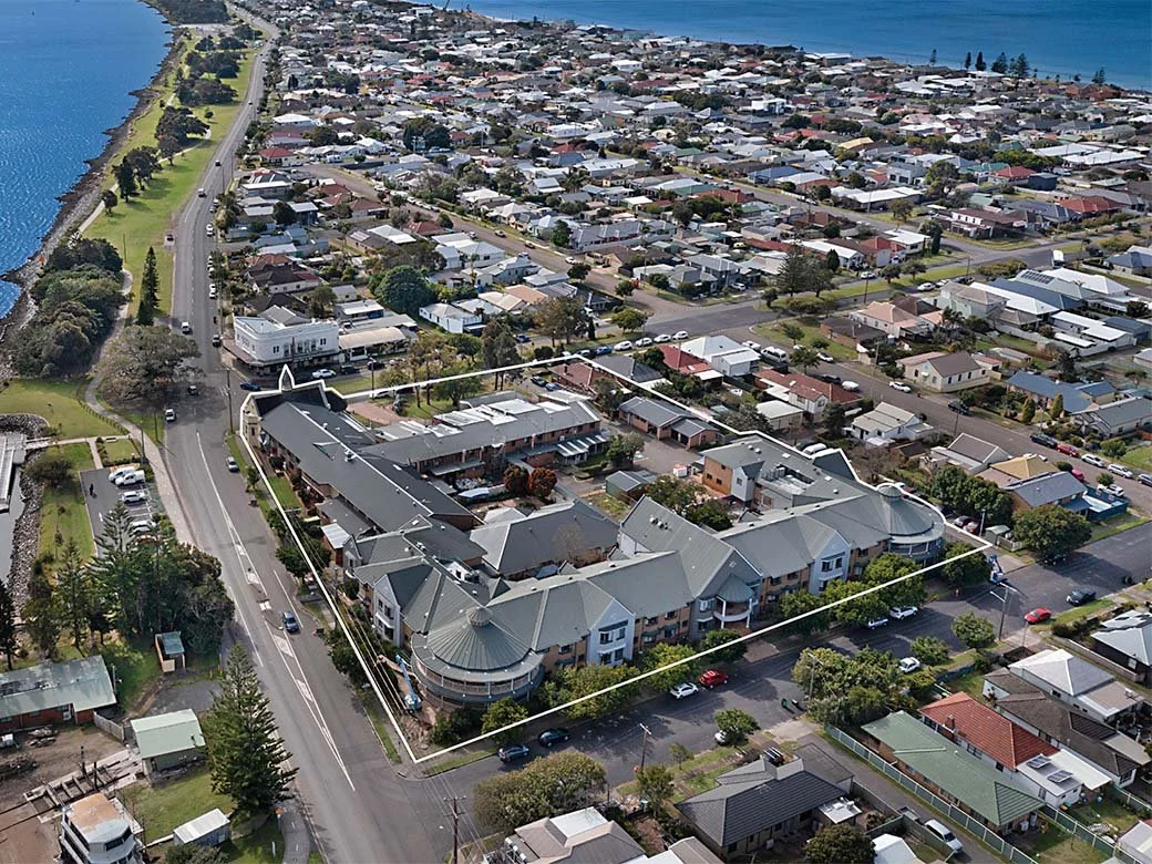 Aerial view of a coastal residential neighborhood with a large complex of apartment buildings outlined with white lines, located near water and surrounded by roads, houses, and greenery.