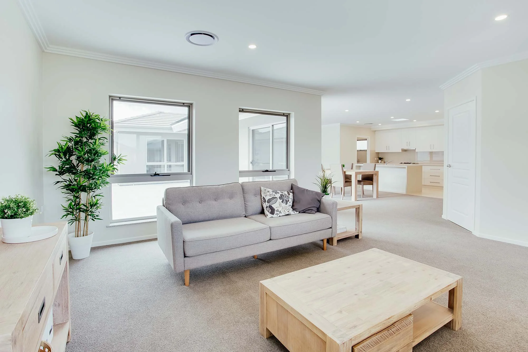 Bright, minimalist living room with light-colored sofa, wooden coffee table, and indoor plants, open to a kitchen and dining area.