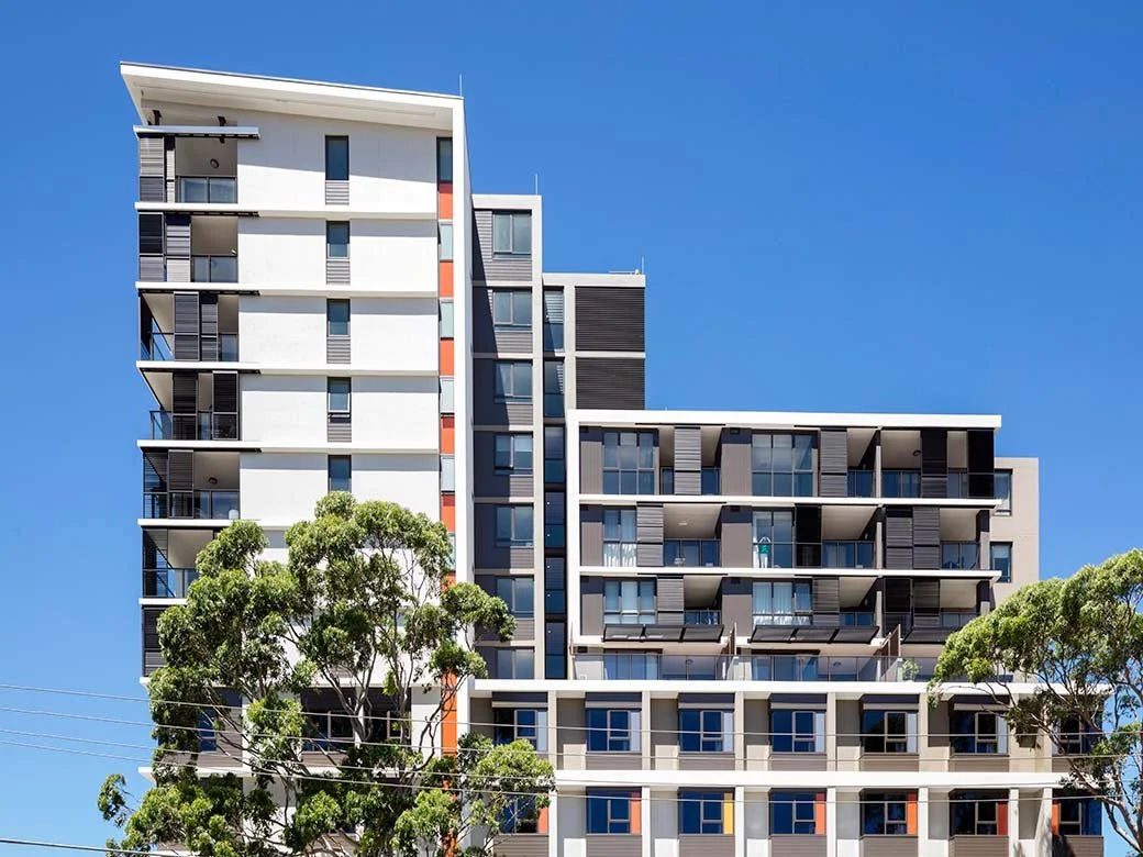 Modern multi-story apartment building with balconies and large windows, set against a clear blue sky, with some green trees in the foreground.