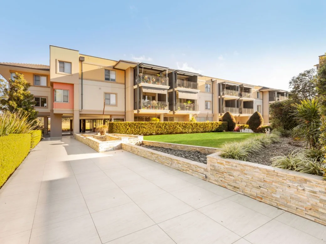 Modern apartment building with landscaped lawn, bushes, and paved walkway under a clear blue sky.