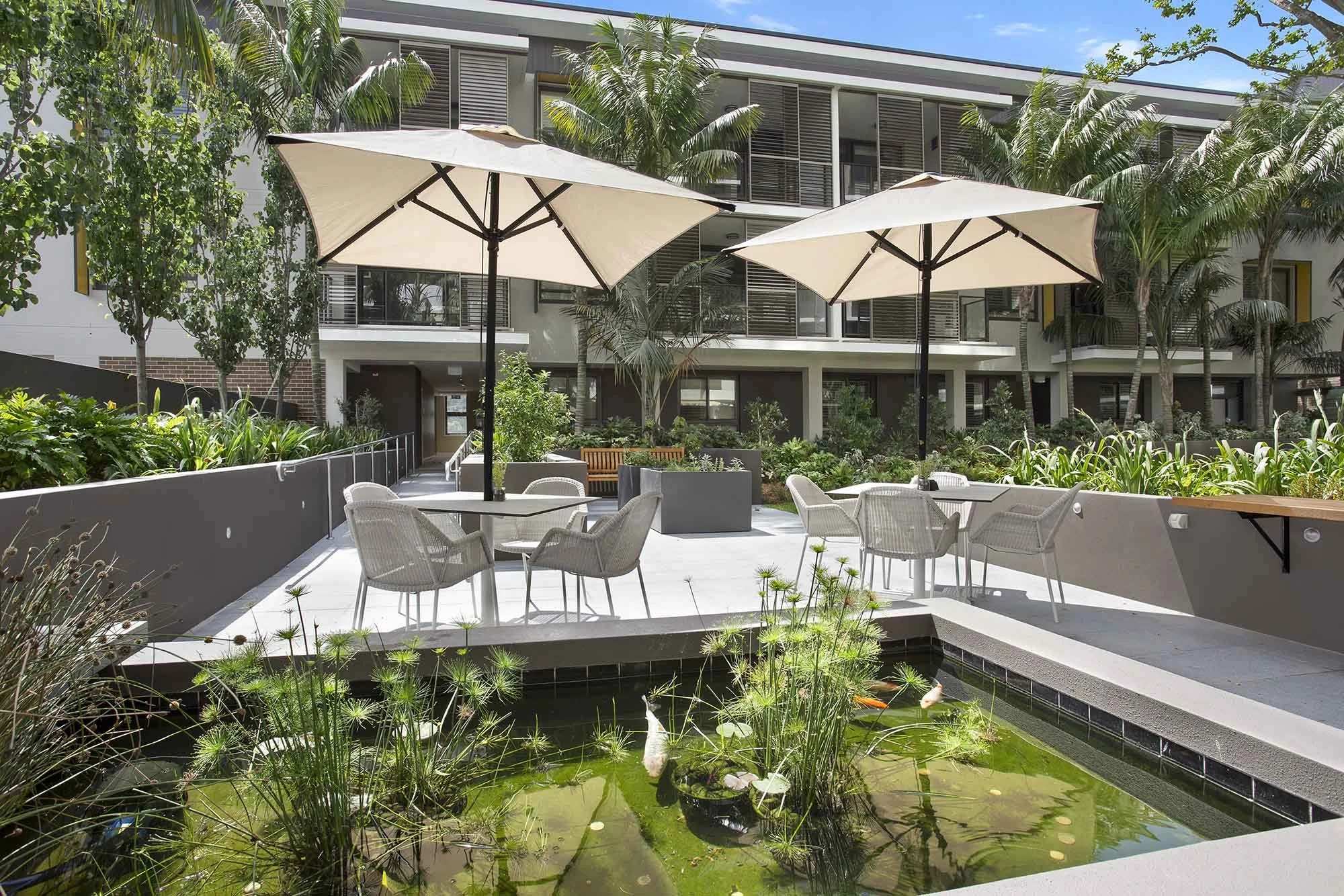 Outdoor patio area with white chairs, tables, and umbrellas, a small pond with plants and fish, surrounded by greenery and modern apartment buildings.