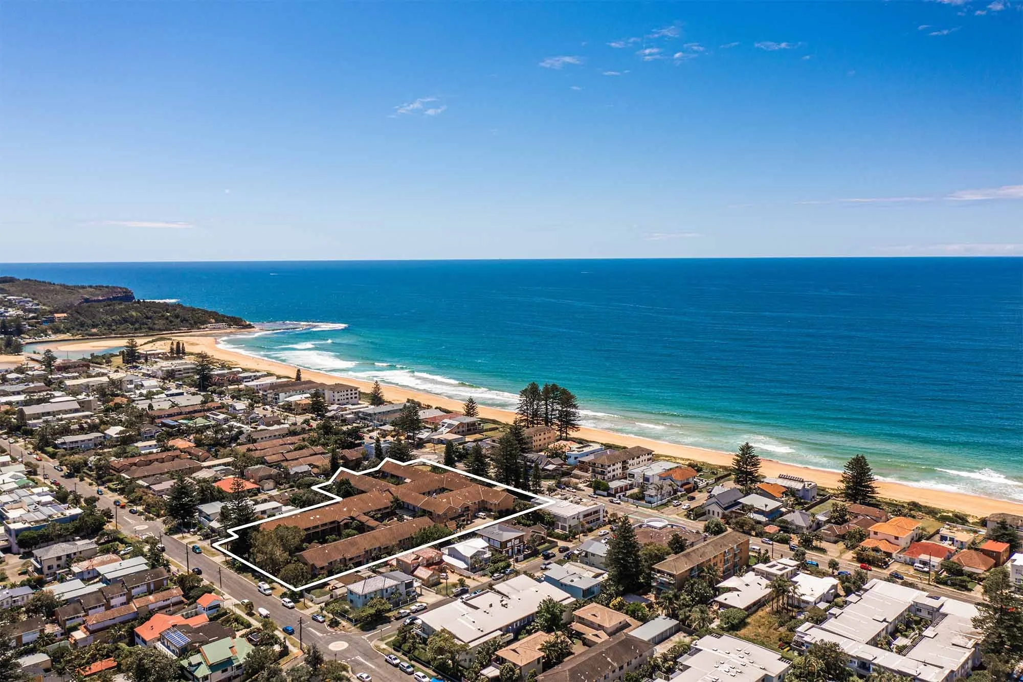 Aerial view of a coastal neighborhood with houses and streets near the beach, featuring a outlined property, with the ocean and sandy beach in the background under a bright blue sky.