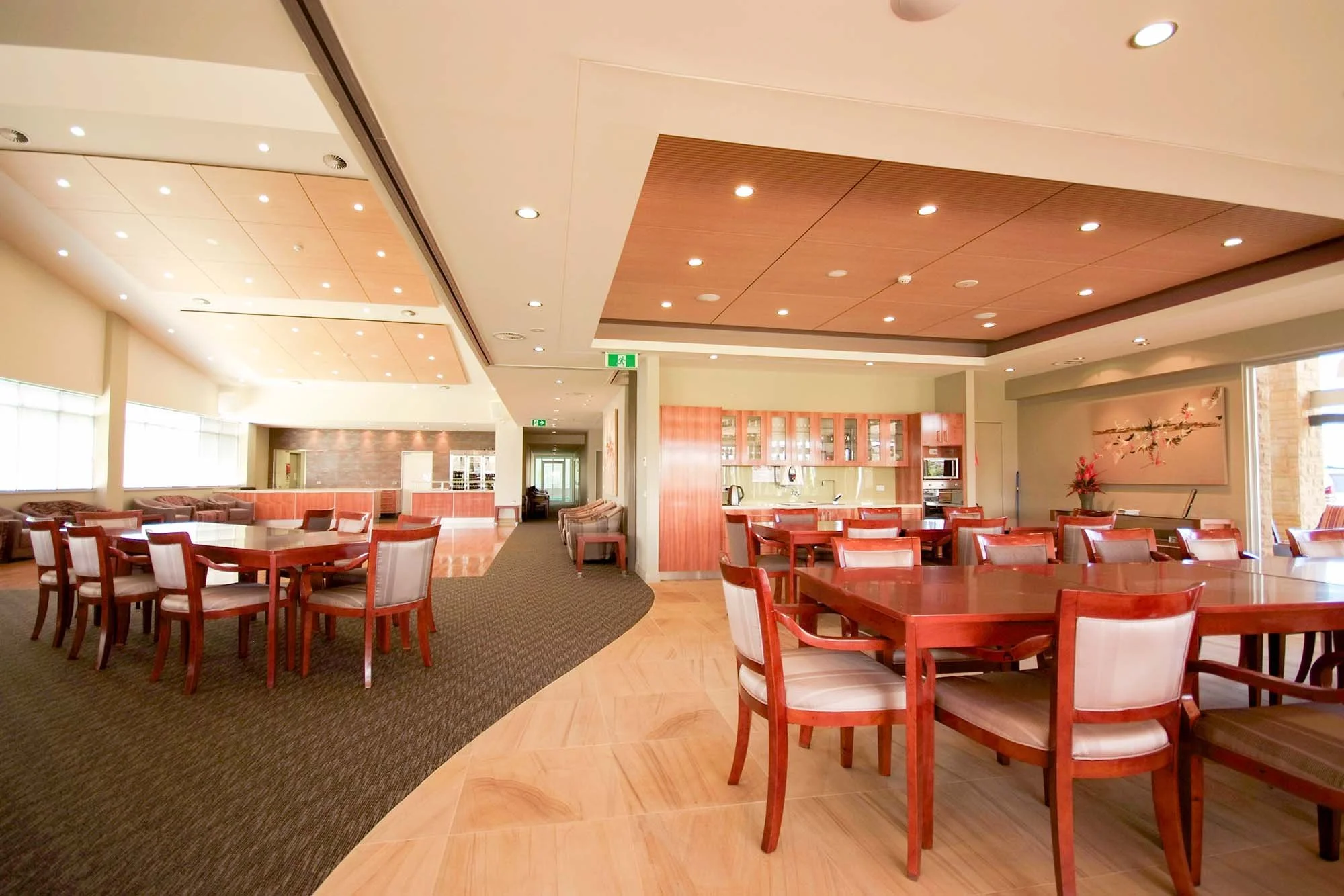  Spacious restaurant with wooden tables and chairs, a breakfast bar area, natural light from large windows, and modern ceiling lighting, decorated with a flower painting and a potted plant.