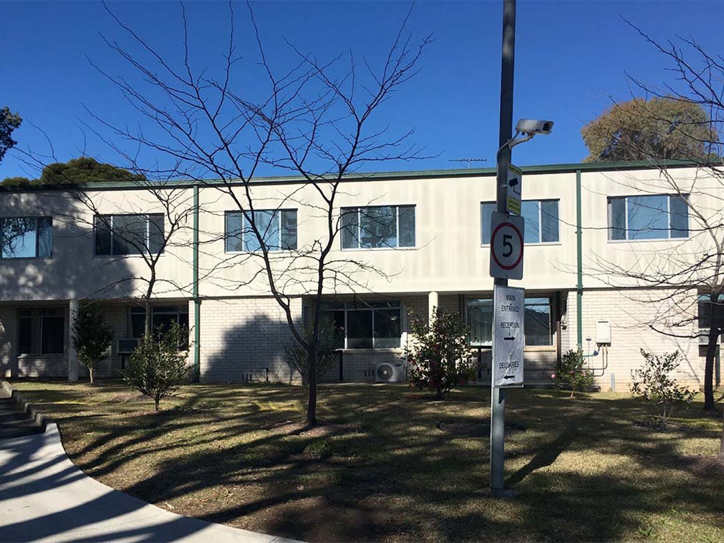 A two-story building with white walls, multiple rectangular windows, and a small landscaped area with trees and bushes in front. A signpost with a speed limit of 5 and directions for main entrance, reception, and deliveries is in the foreground, under a clear blue sky.