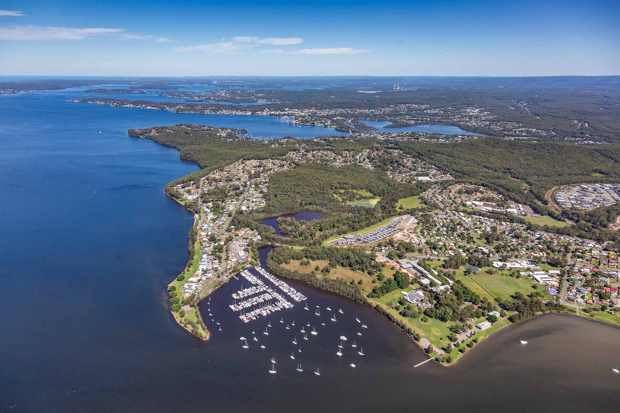 Aerial view of a coastal town with a marina filled with sailboats, surrounded by green parks, residential areas, and large bodies of water under a clear blue sky.