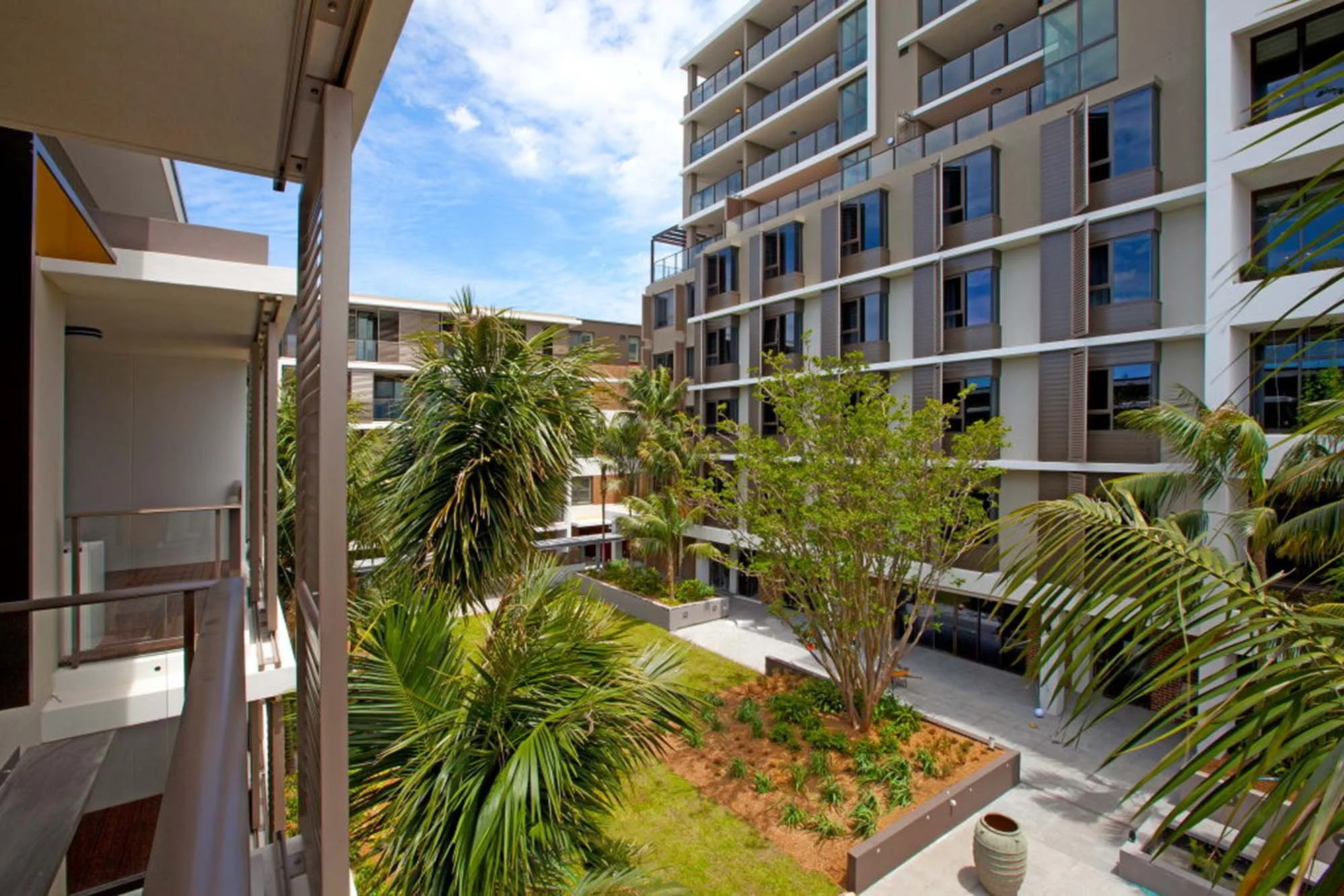 View of an apartment complex courtyard with palm trees, greenery, and modern buildings under a blue sky.