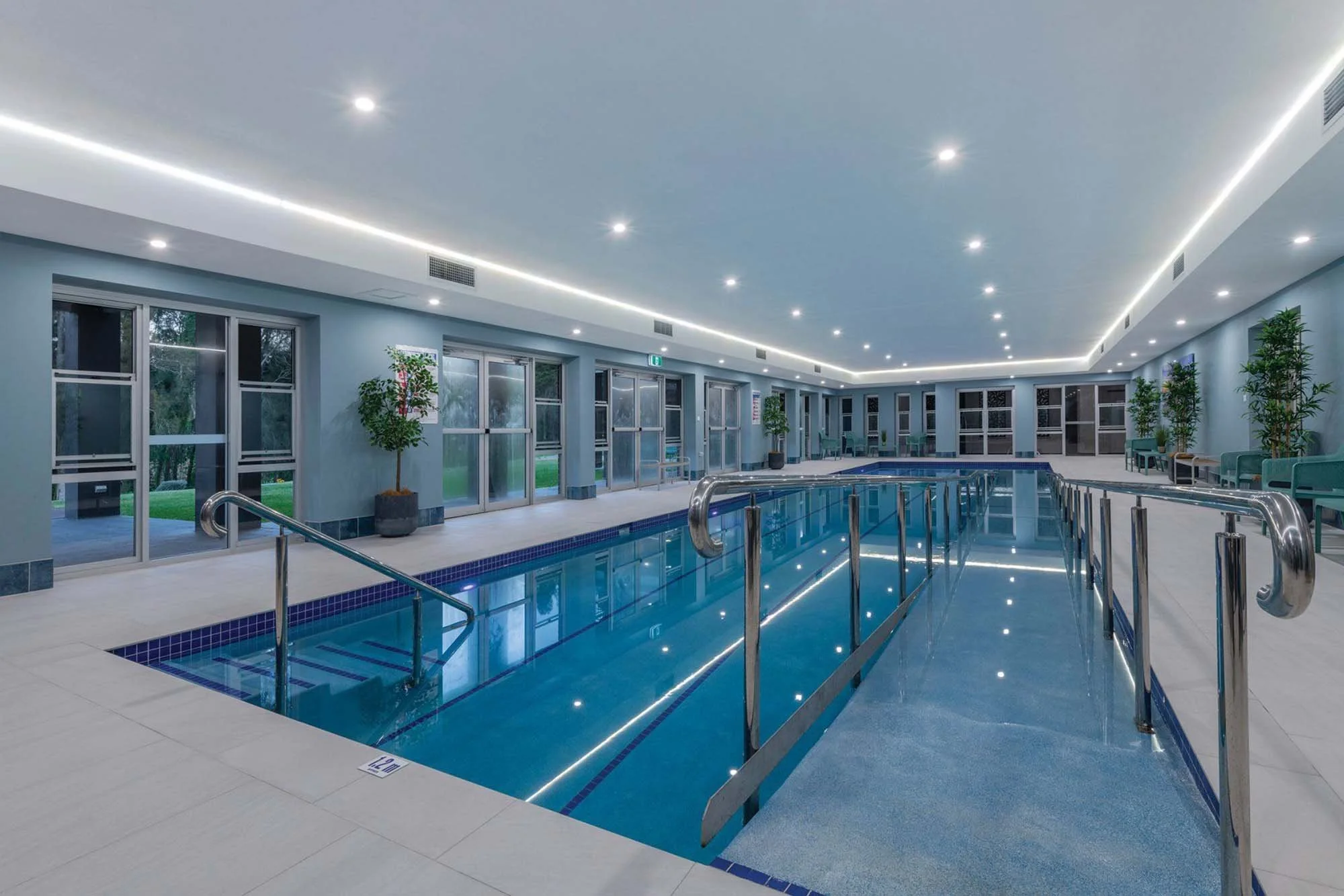 Indoor swimming pool with a ramp for accessibility, surrounded by potted plants, windows, and seating areas, with bright ceiling lights