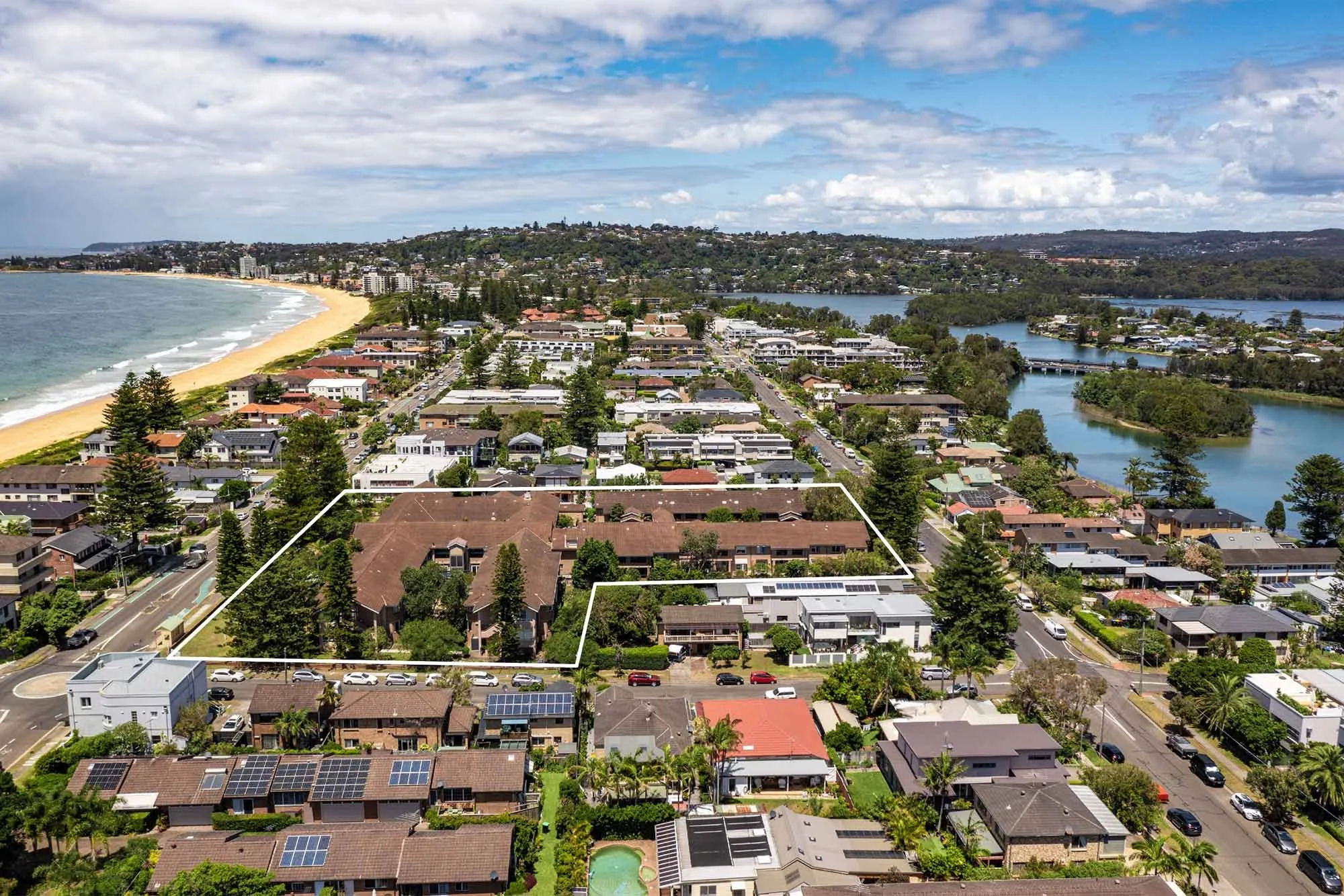 Aerial view of a residential area near a beach with houses, trees, a canal, and a sandy shoreline under a partly cloudy sky.