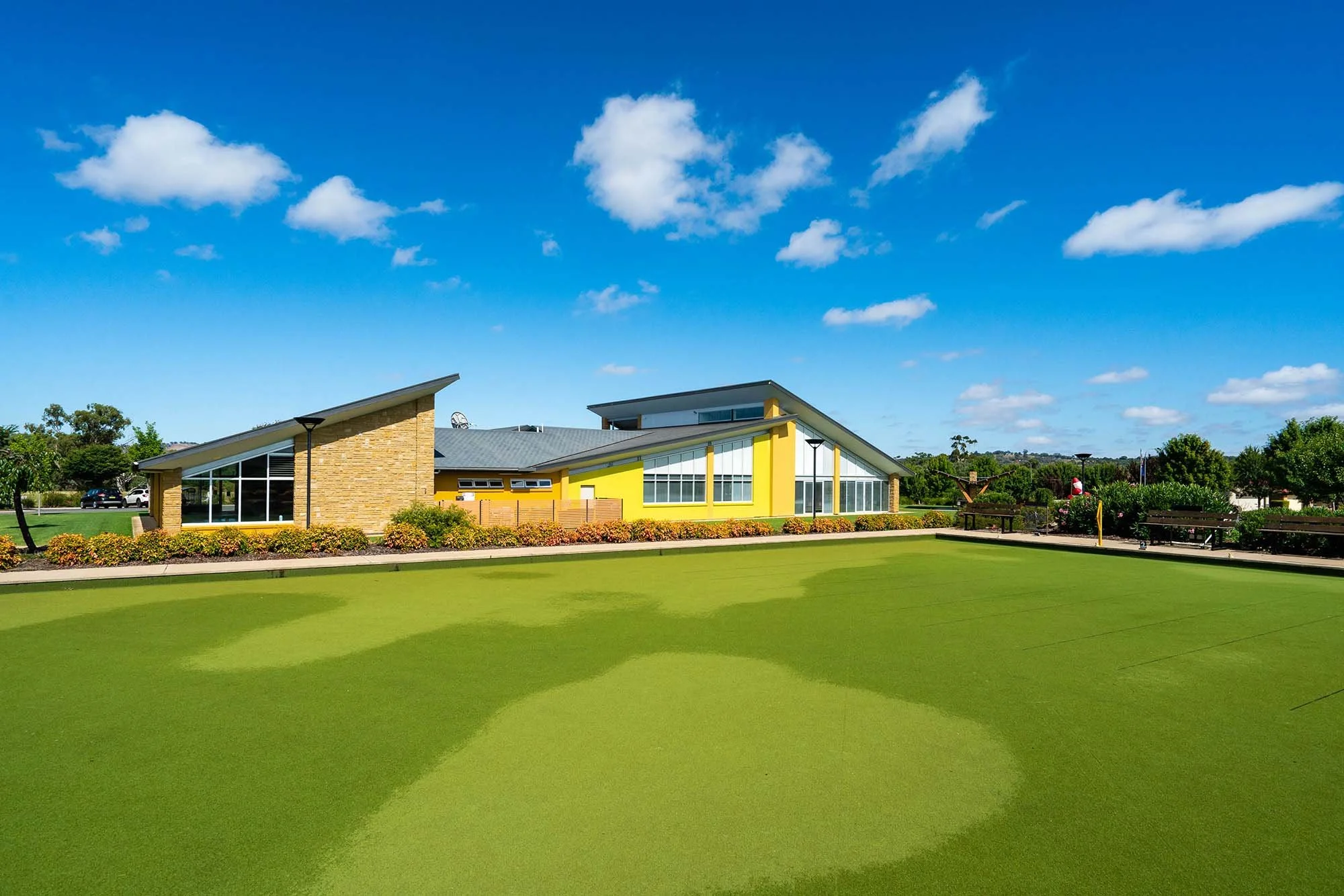 Modern building with sloped roof and large windows, surrounded by vibrant greenery and benches, under a partly cloudy blue sky.