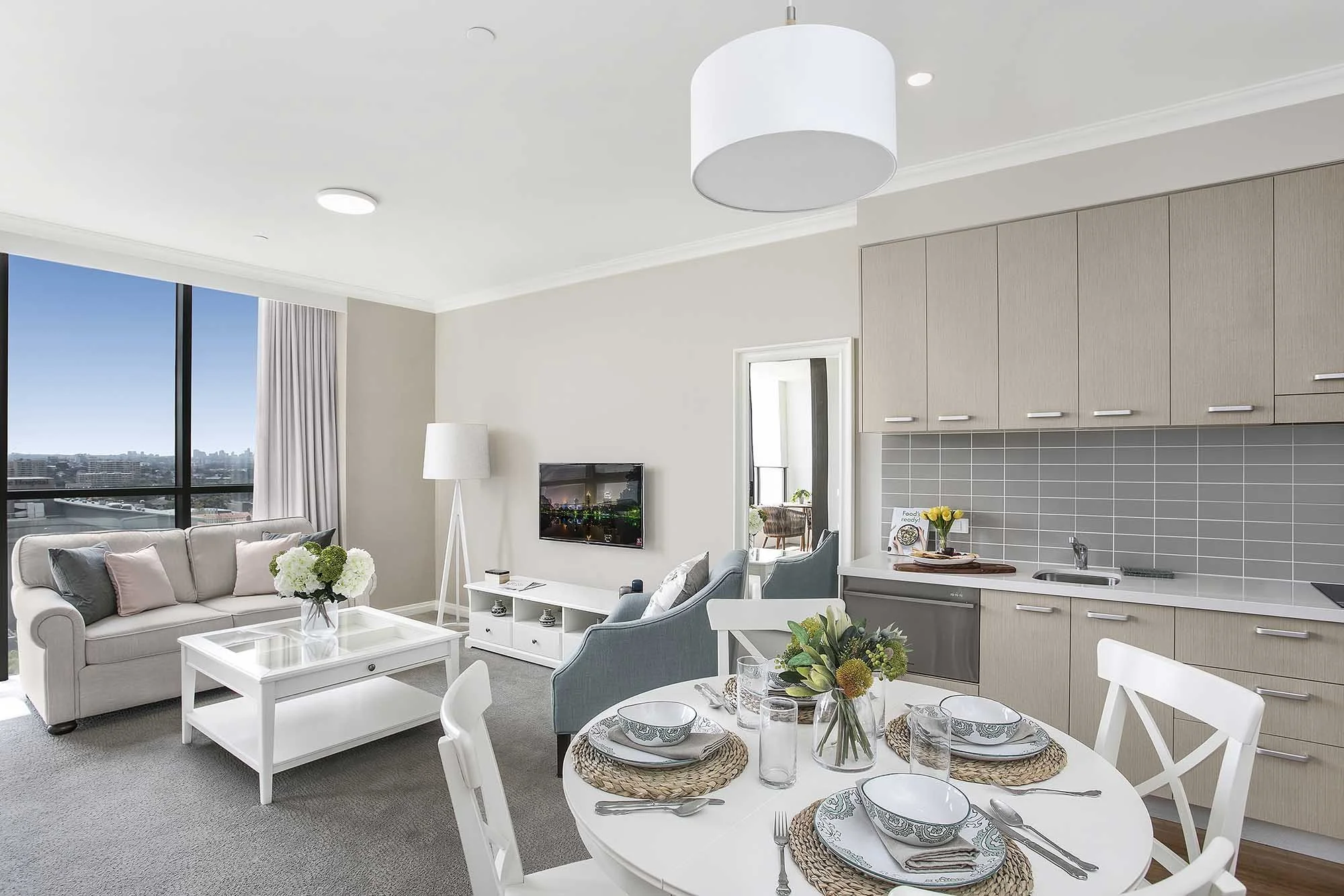 Bright living room and dining area with white furniture, a round dining table set with plates, glasses, and cutlery, a sofa with cushions, a TV mounted on the wall, and a large window with city view.