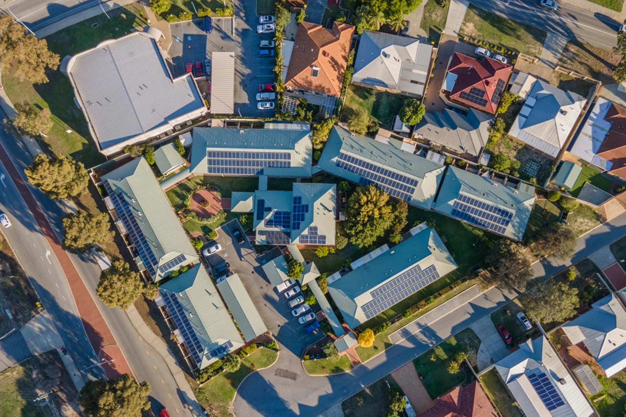 An aerial view of a residential neighborhood with houses featuring solar panels on their roofs, surrounded by trees, lawns, parking areas, and streets.