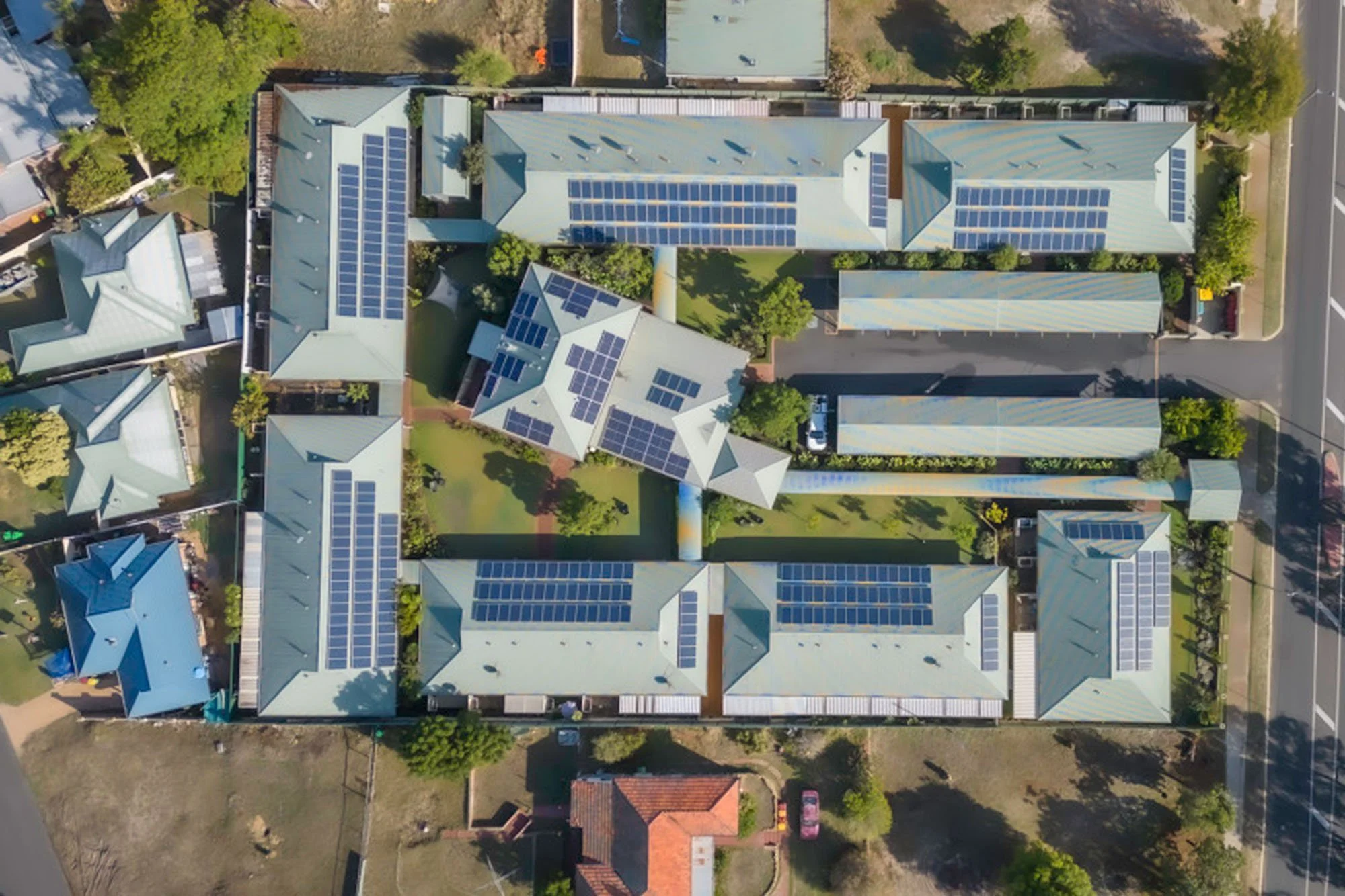 Aerial view of multiple buildings with solar panels on roofs, surrounded by green trees, paved pathways, and parking areas.
