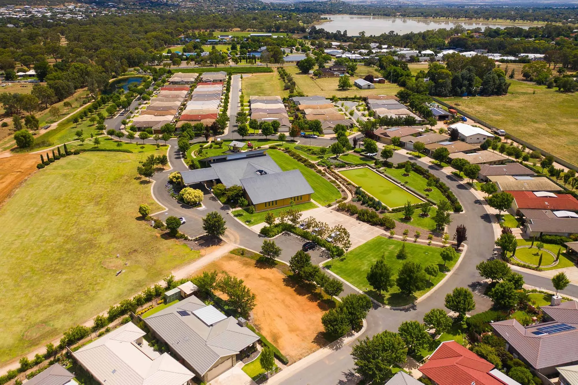 Aerial view of a suburban neighborhood with houses, green lawns, and a pond, with a large green space, tennis court, and lake in the background.