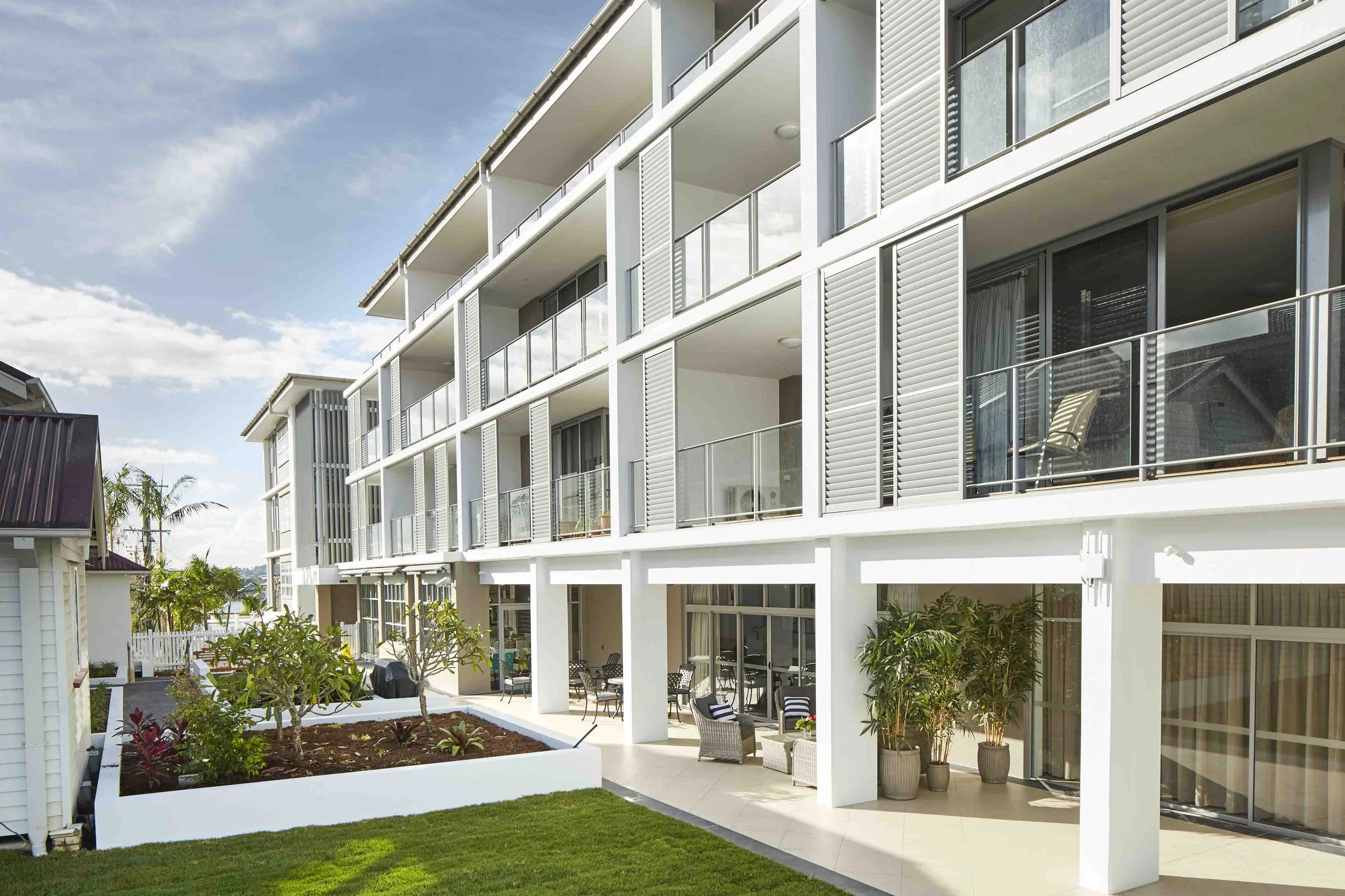 Exterior view of a modern apartment building with balconies, large windows, and a ground-level patio with outdoor furniture, plants, and a small garden.