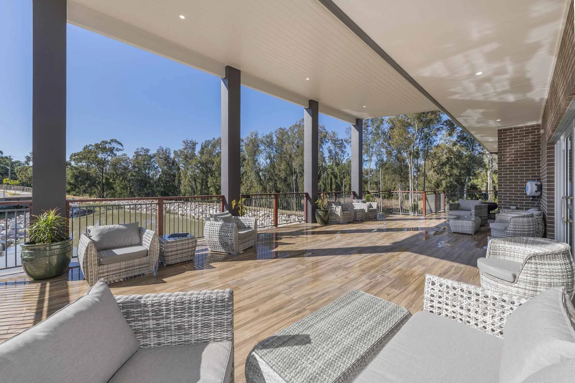 Spacious outdoor balcony with wicker furniture, potted plants, and a wooden floor, overlooking a river and trees on a sunny day.