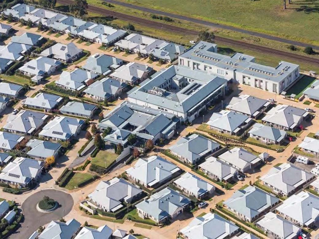 Aerial view of a suburban neighborhood with single-family houses and a large school or commercial building, surrounded by green lawns and streets.