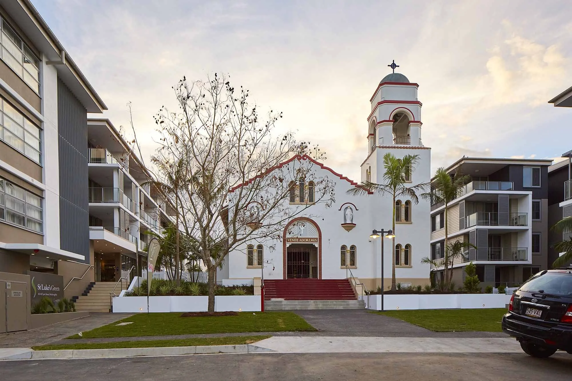 A church surrounded by modern residential buildings with a tree in front, during sunset.