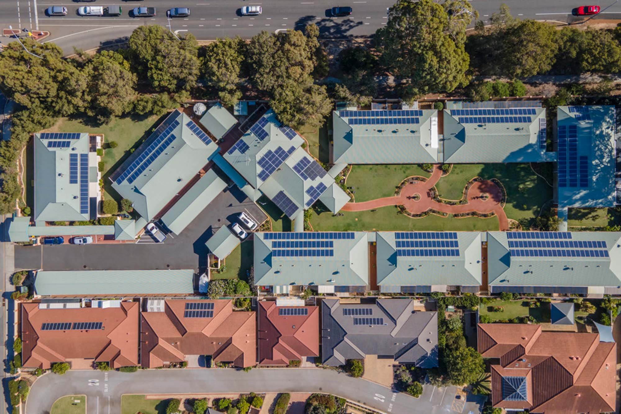 Aerial view of residential buildings with solar panels on roofs, a small courtyard with pathways and greenery, and a parking lot with cars at the top of the image.