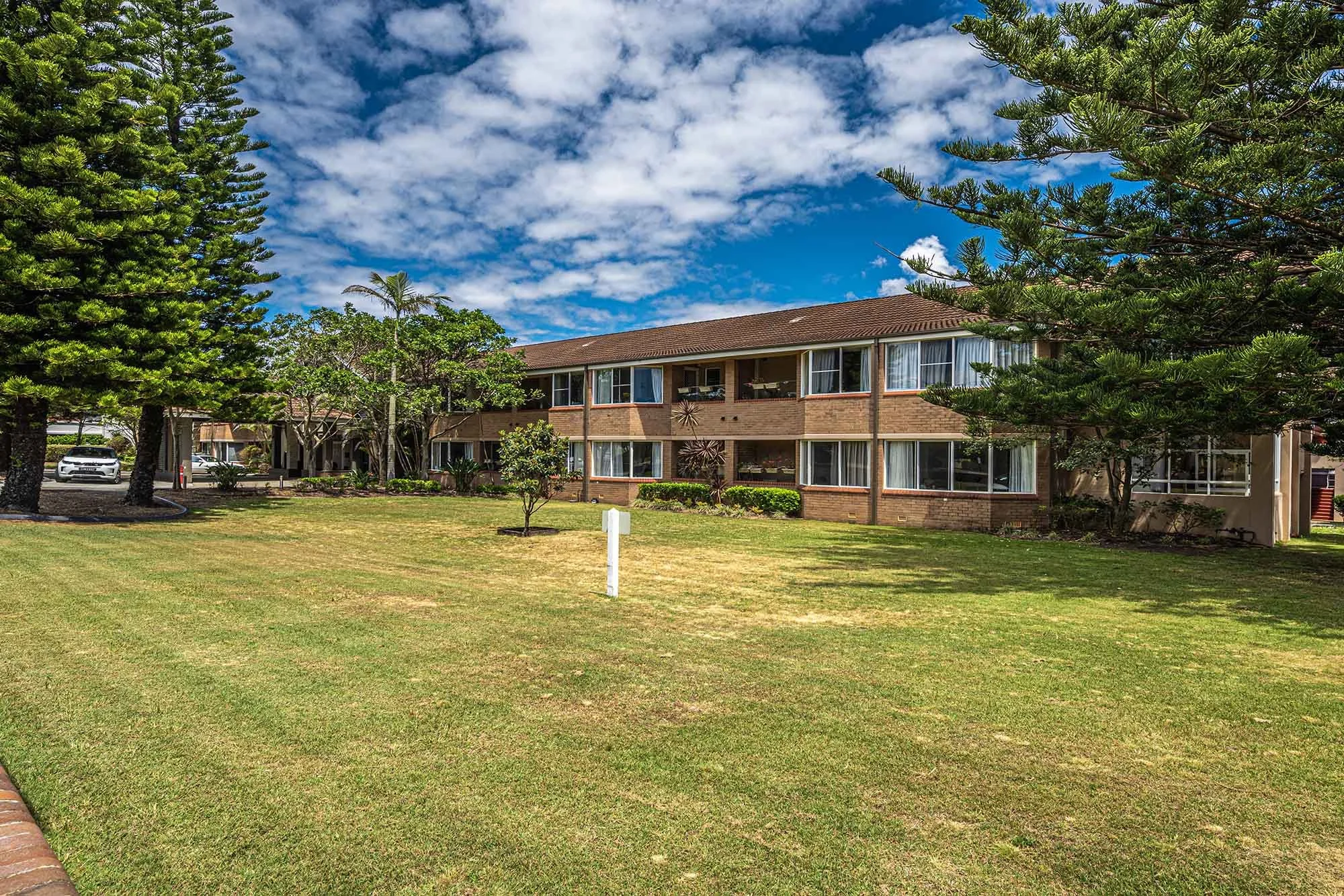A two-story apartment building with large windows, surrounded by green trees and a well-maintained lawn. The sky is partly cloudy with blue and white clouds.