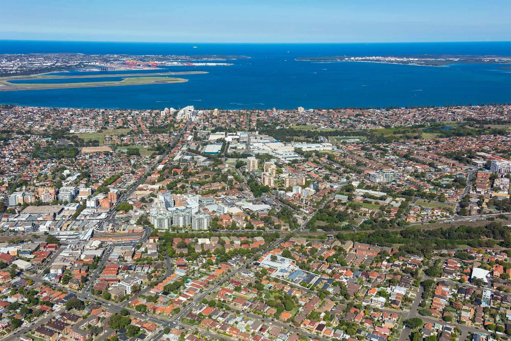 Aerial view of a coastal city with residential, commercial, and industrial buildings, green parks, and a large body of water in the background.