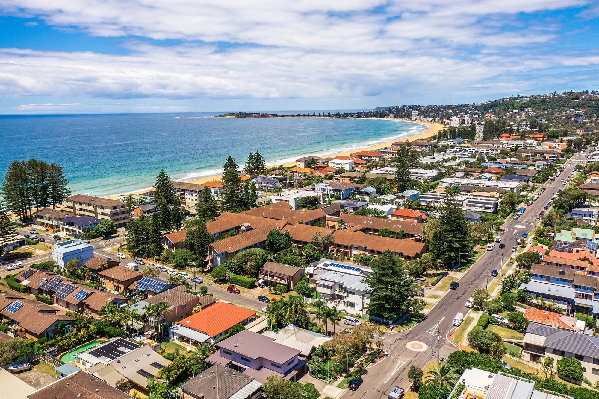 Aerial view of a coastal city with residential houses, streets, and a beach with white sand and turquoise water under a partly cloudy sky.