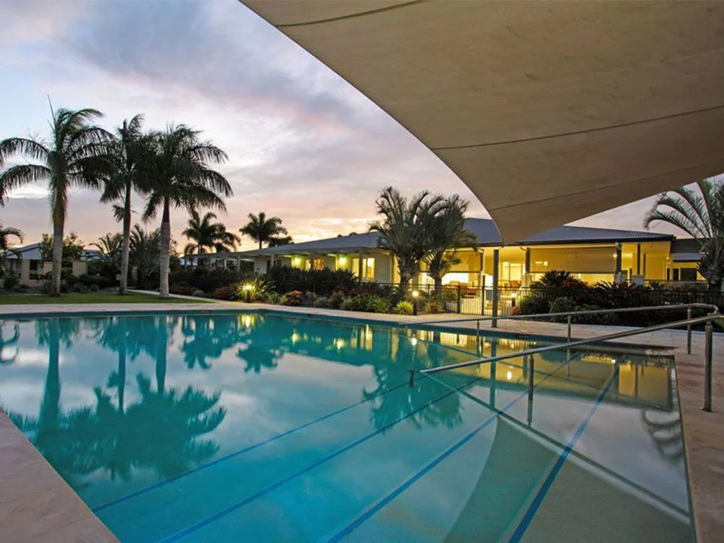 Swimming pool in a resort with palm trees, building with lights, and a cloudy sky at sunset.