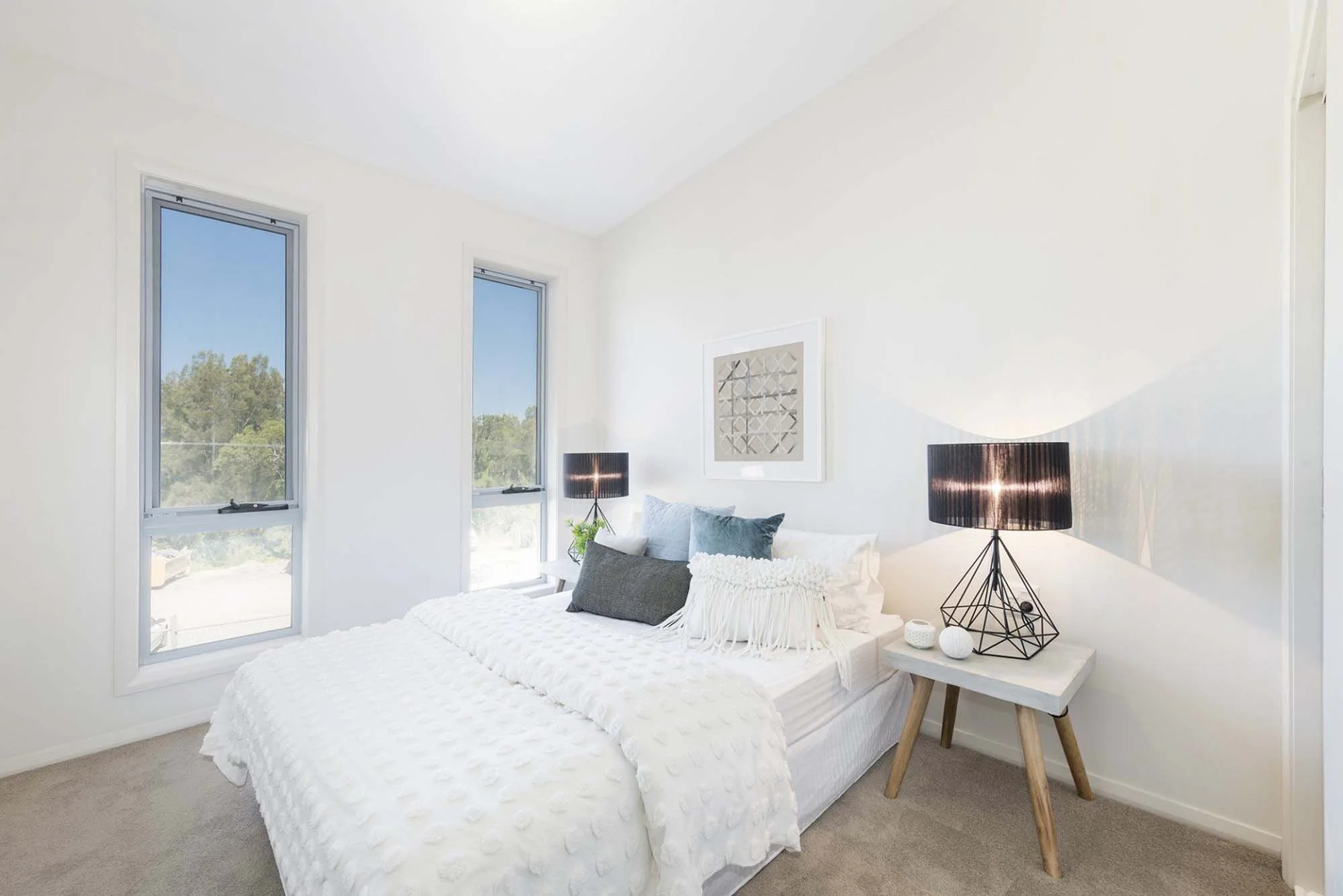 Bright white bedroom with a bed covered in a textured white blanket, multiple gray and white pillows, two black table lamps on a light-colored wooden side table, a framed geometric artwork above the bed, and three tall windows with views of green tre