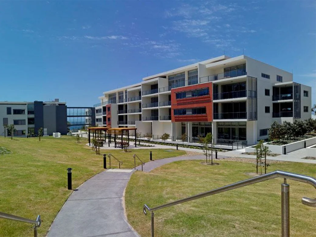 Modern multi-story apartment buildings with balconies in a landscaped area with a walking path and small trees under a clear blue sky.