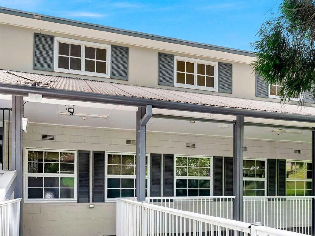 Exterior view of a multi-story building with white walls, dark gray window shutters, and a covered balcony with white railings, under a blue sky with some clouds.