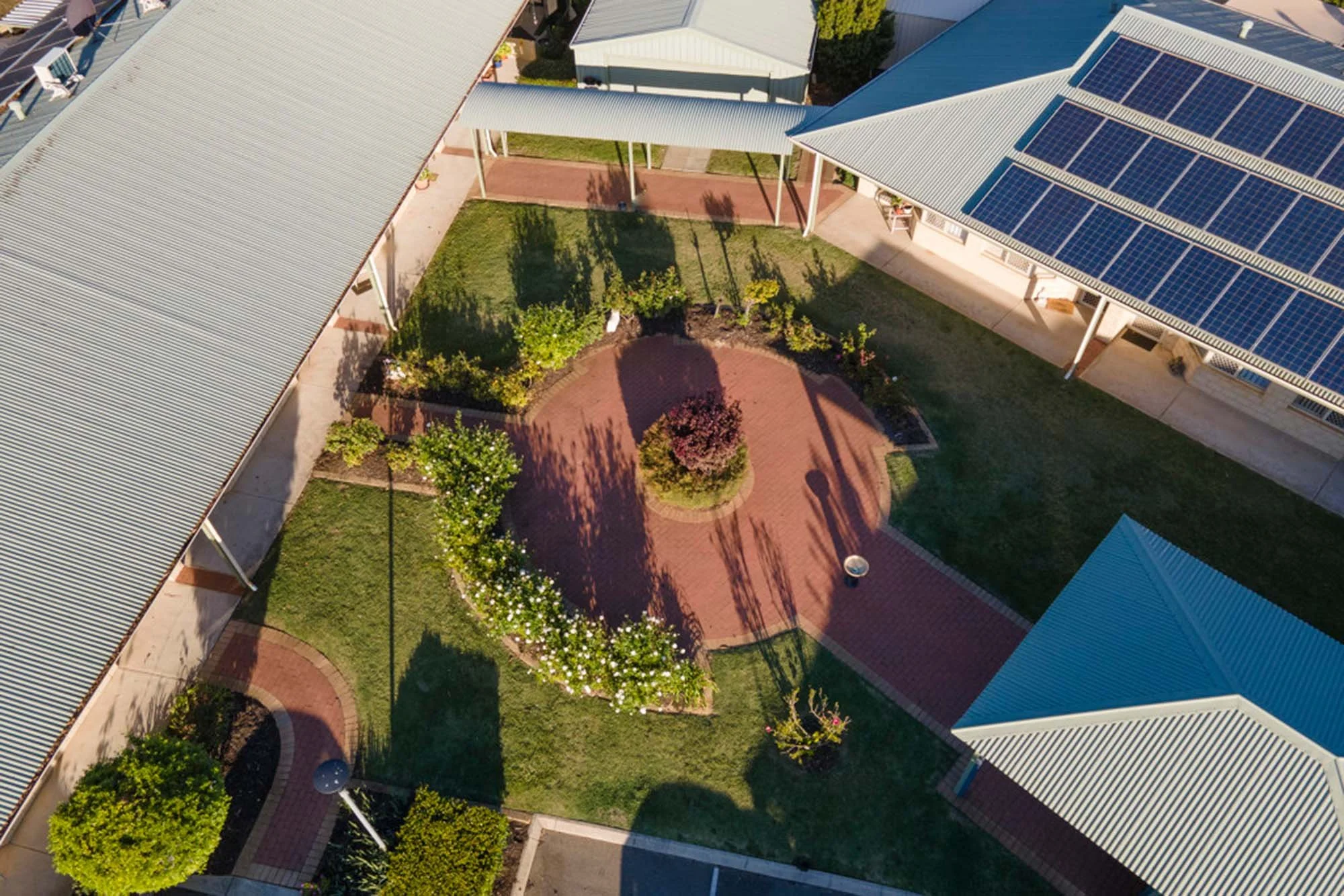 An aerial view of a small courtyard with a circular brick pathway, surrounded by green grass, shrubs, and trees. There are buildings with metal roofs, some covered with solar panels, casting shadows on the ground.