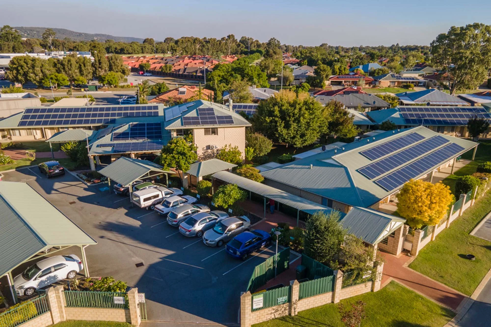 Residential community with multiple buildings covered in solar panels on roofs, parking lot with cars, surrounded by trees and greenery.