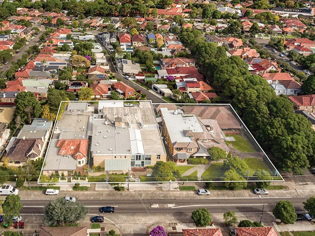Aerial view of a residential neighborhood with houses, streets, green trees, and a marked property boundary around a large complex of buildings.