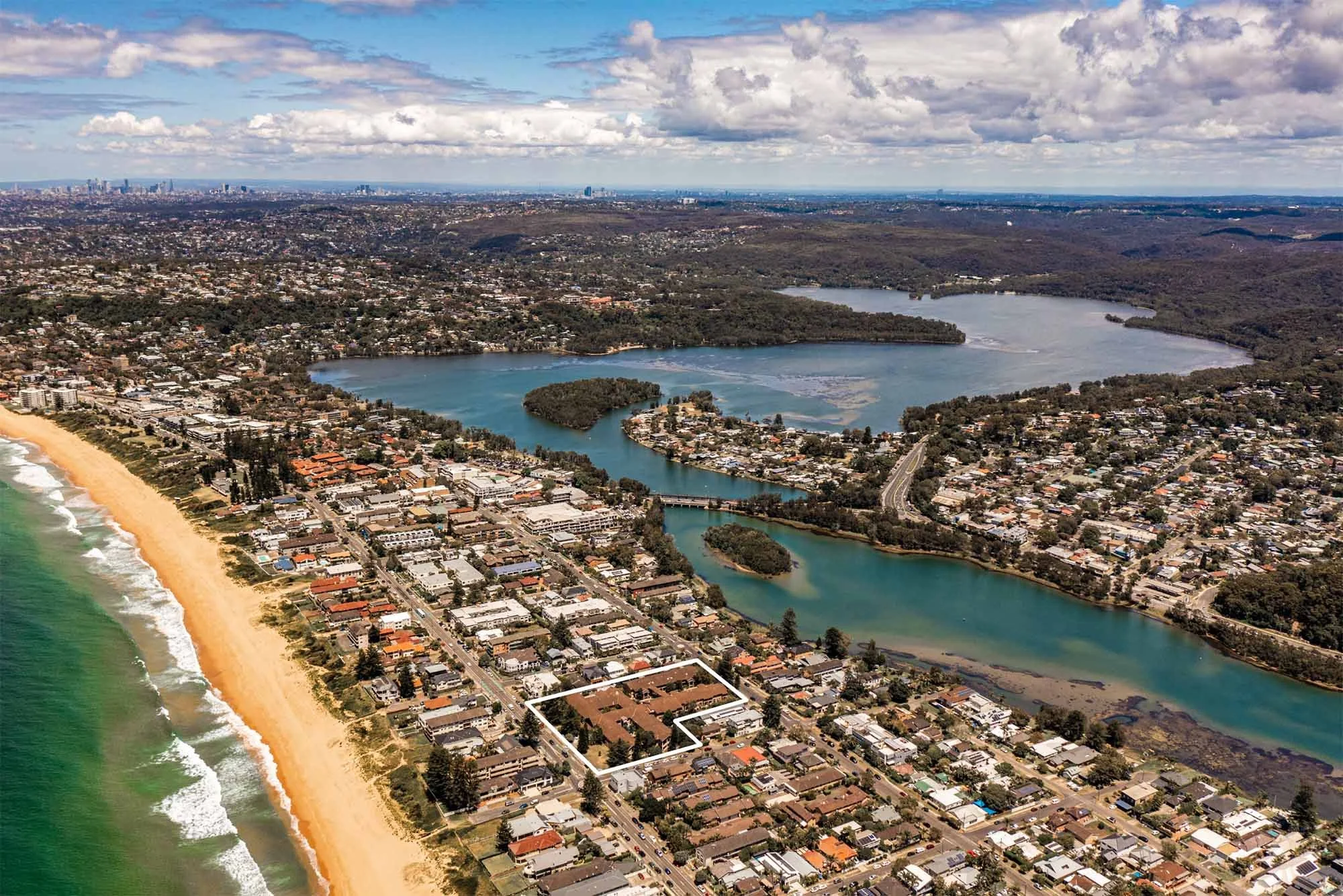 Aerial view of a coastal city with a beach, ocean on the left, and waterways with parks and residential areas on the right.