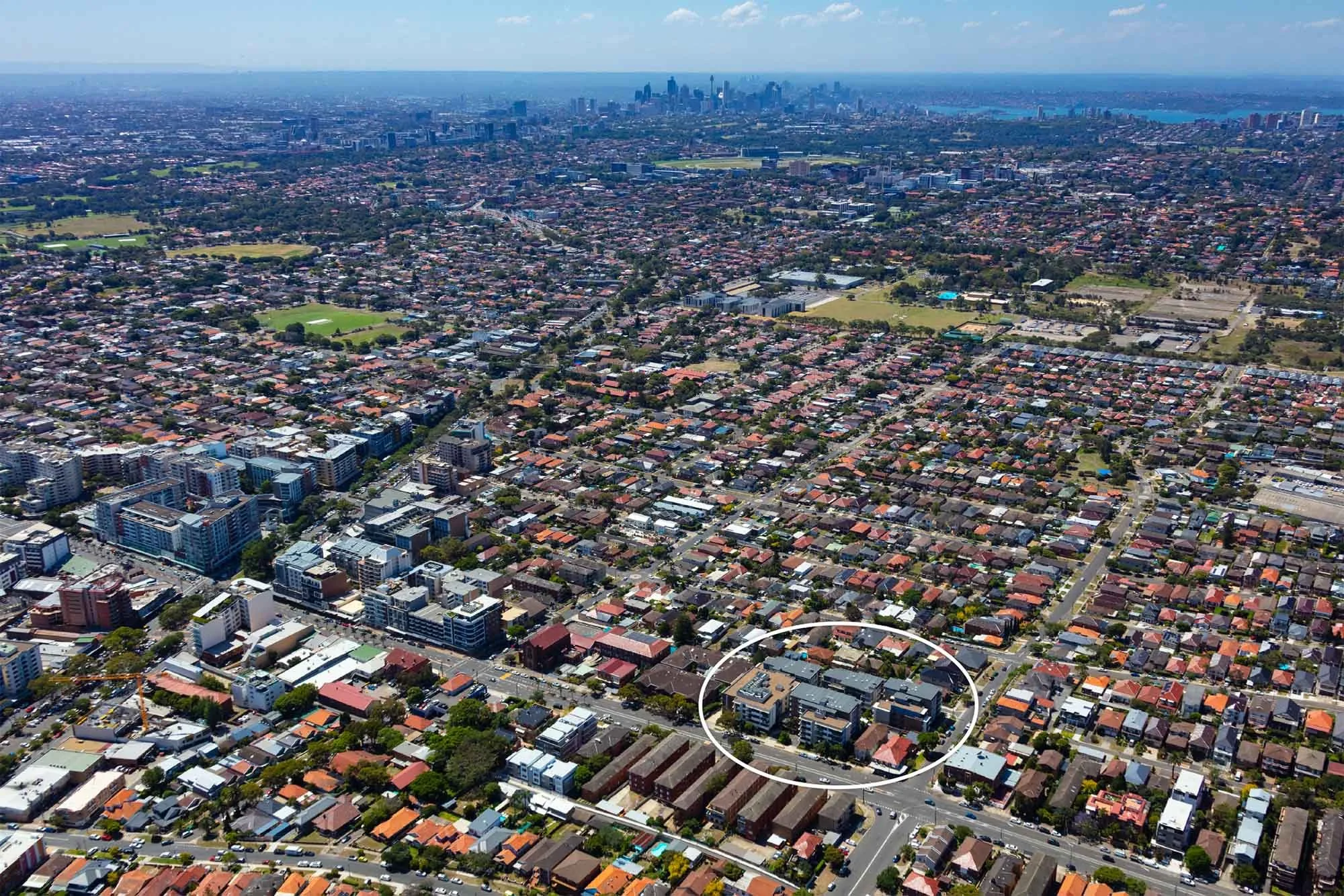 Aerial view of a cityscape showing a densely populated area with residential houses and some commercial buildings, with a downtown skyline and water in the distance.