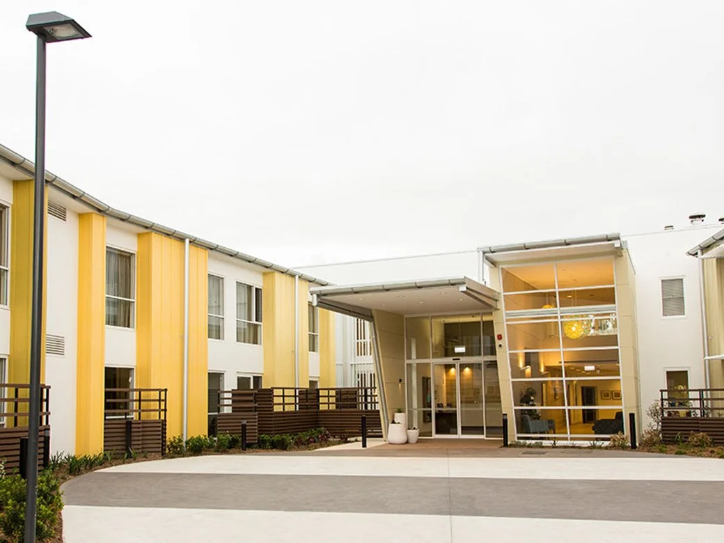 Exterior view of a modern apartment building with a glass front entrance, yellow and white walls, and landscaped surroundings.