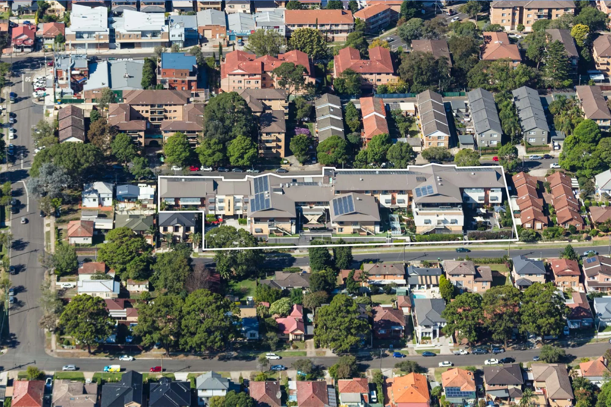 Aerial view of a residential neighborhood with a large property outlined in white, featuring multiple buildings, trees, and solar panels on roofs.