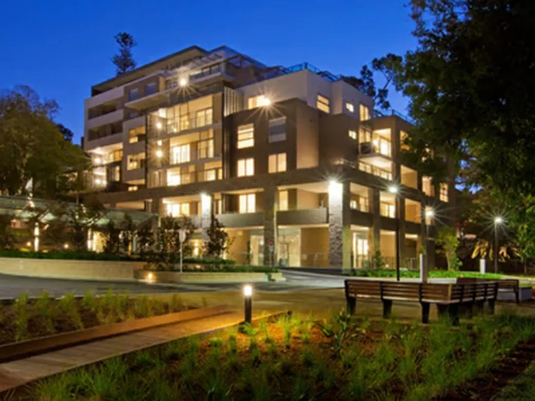 Modern multi-story apartment building illuminated at night, surrounded by landscaping and outdoor lighting.