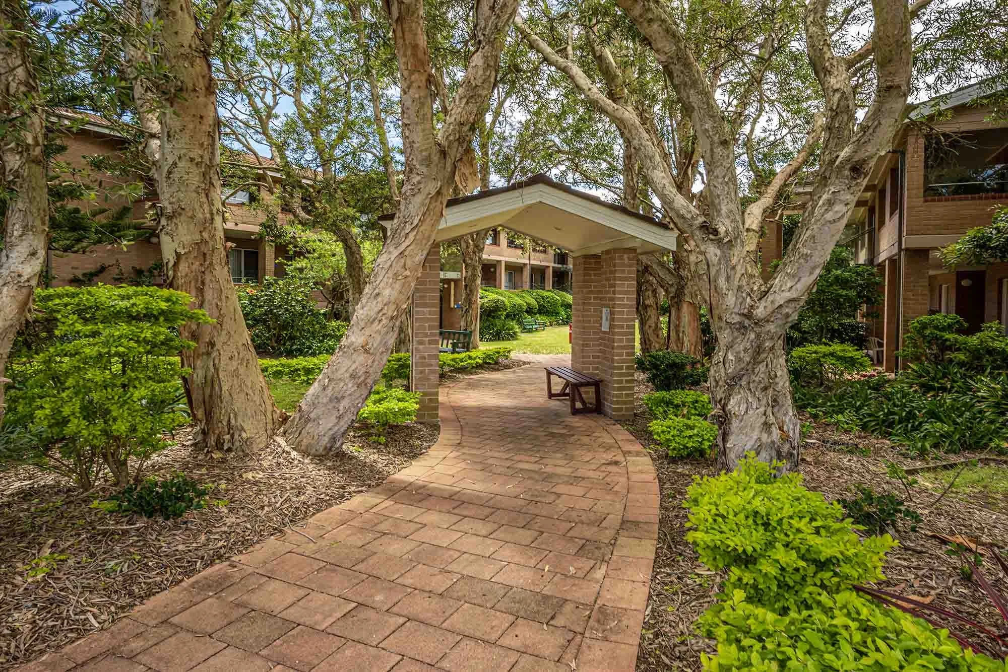 A brick pathway leads through a landscaped garden area with green bushes and mature trees, toward a brick pavilion with a sloped roof in a residential complex.