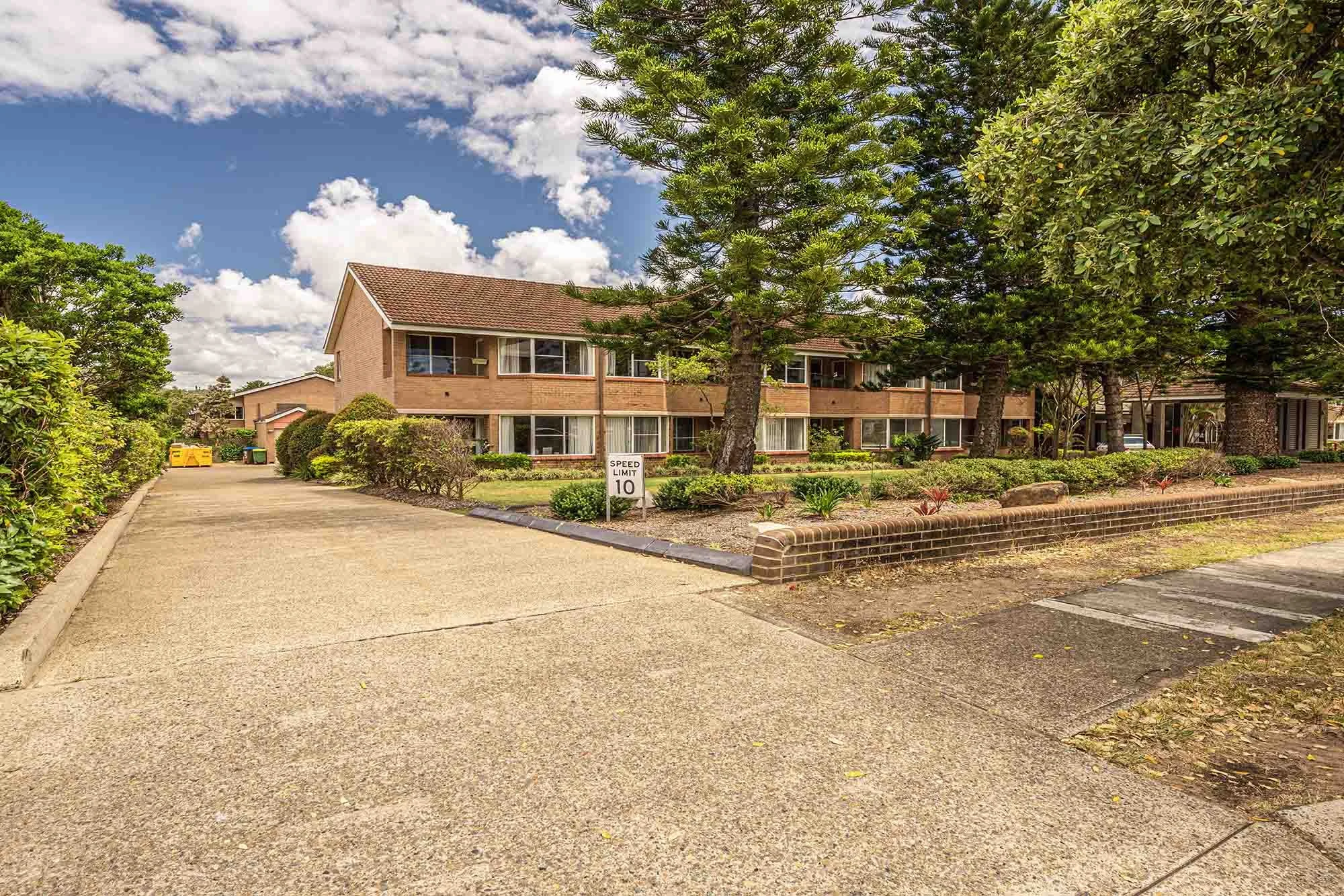 Image of a suburban apartment complex with trees, bushes, a driveway, and a speed limit sign showing 10 mph under a partly cloudy sky.