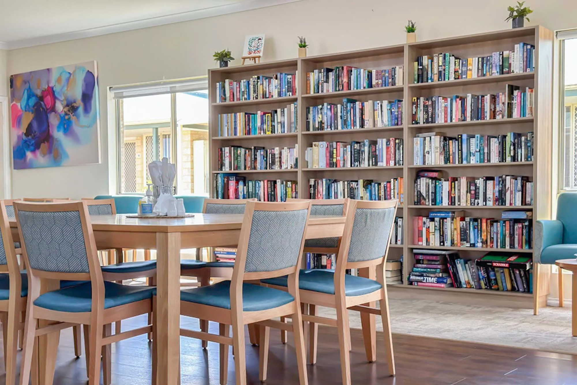 A dining area with a round wooden table and eight wooden chairs with blue cushions, a large bookshelf filled with books, a window, an abstract painting on the wall, and decorative plants on top of the bookshelf.