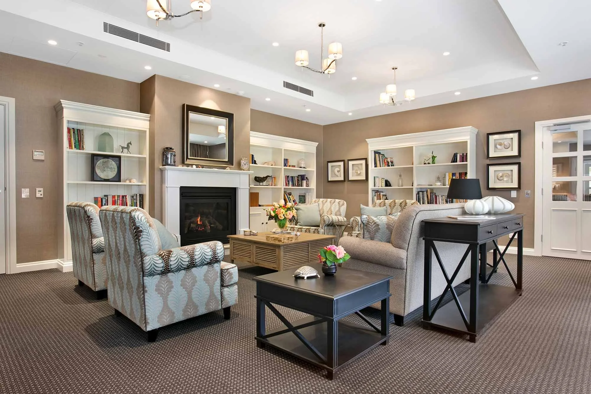 Living room with beige walls, white bookshelves, and a fireplace. Furnished with patterned armchairs, a sofa, black side tables, a beige coffee table, and decorative items including flowers and a mirror.