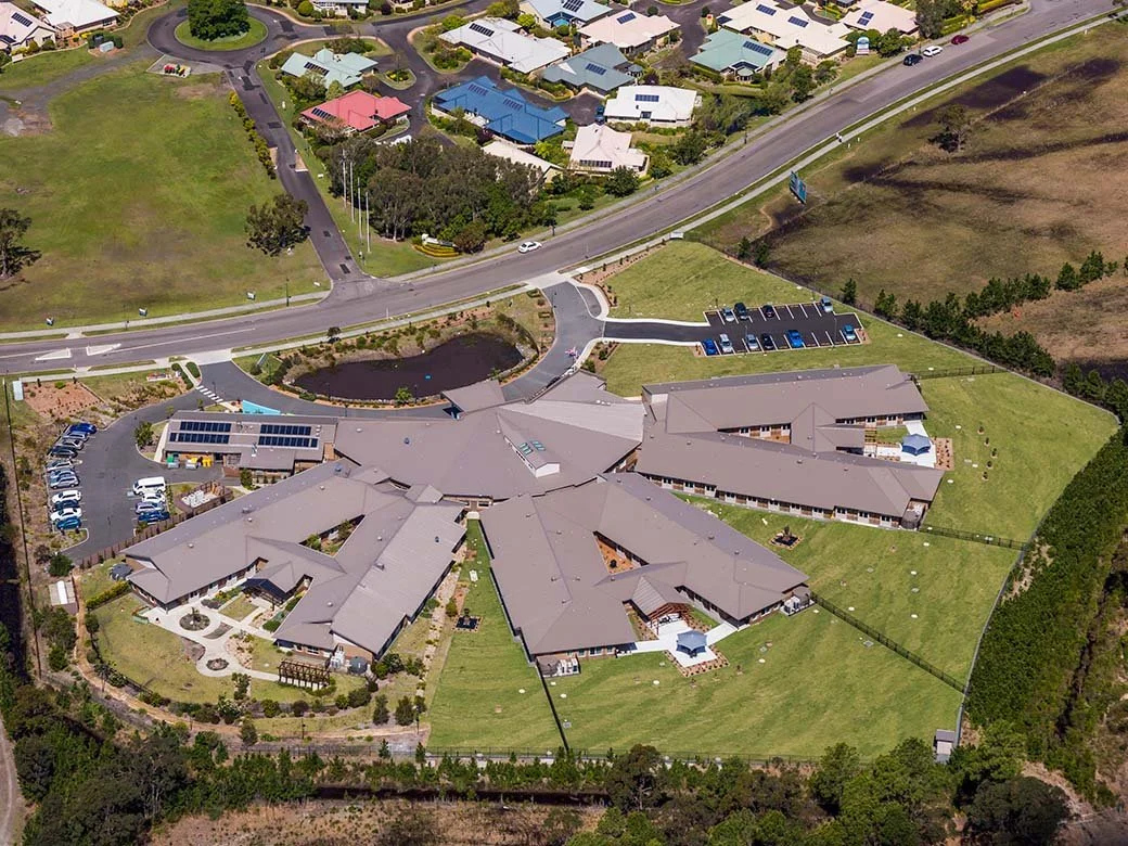 An aerial view of a large residential complex with multiple building wings, parking lots, green lawns, and surrounding roads.