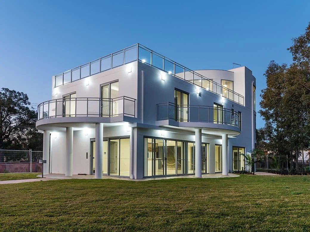 Modern white three-story house with large glass windows and balconies, illuminated at dusk, surrounded by green lawn and trees.