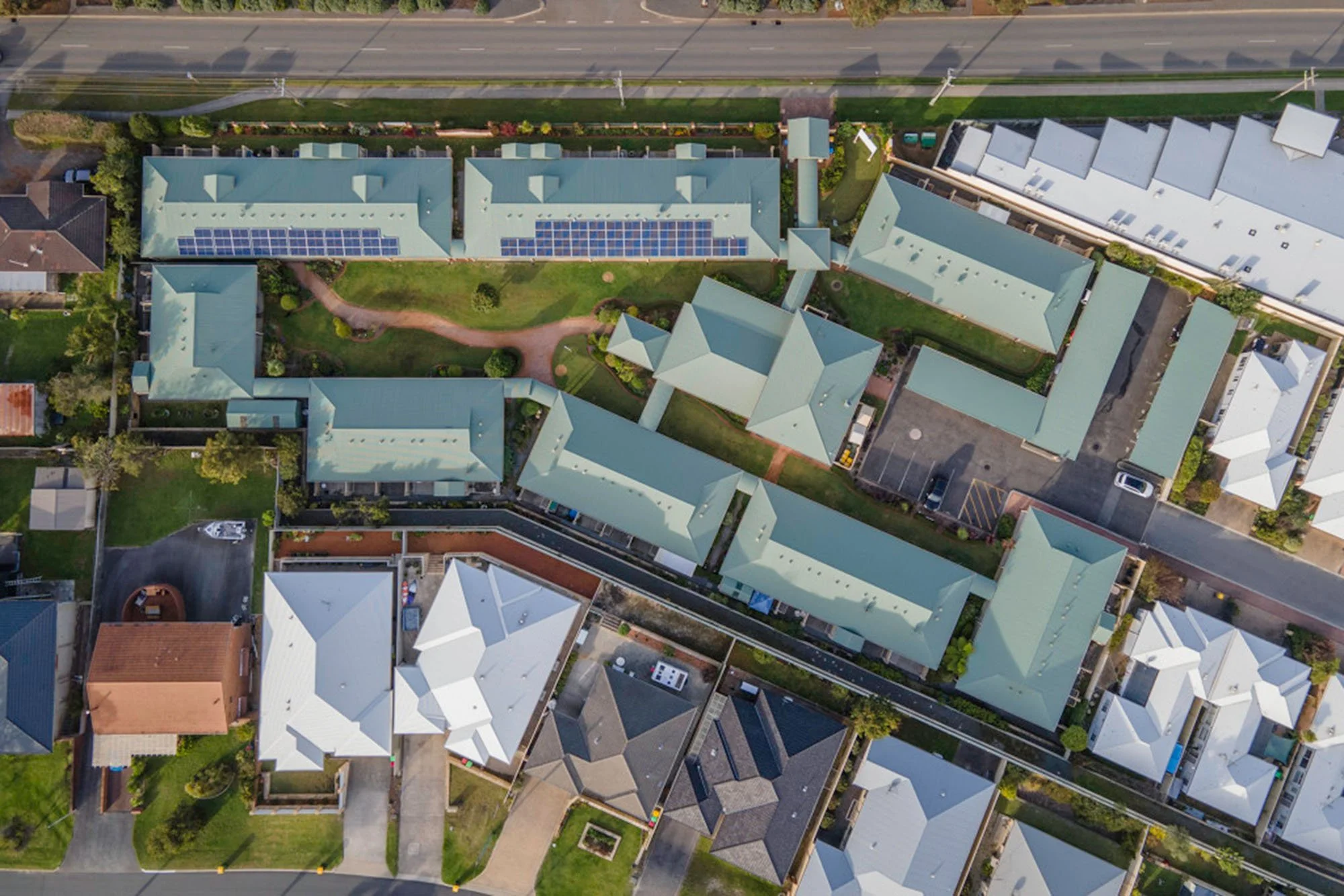 Aerial view of a residential area with multiple houses featuring light-colored roofs, surrounded by green lawns, streets, and some parking lots.