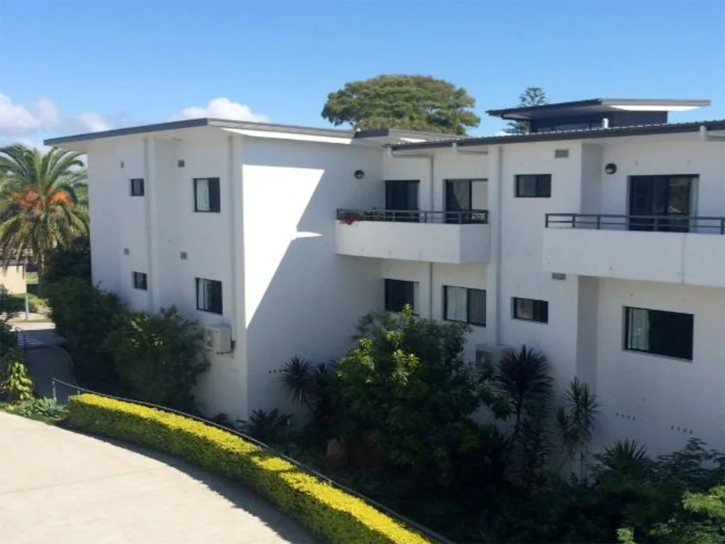 Modern white multifamily apartment building with balconies, surrounded by greenery and palm trees, under a blue sky.