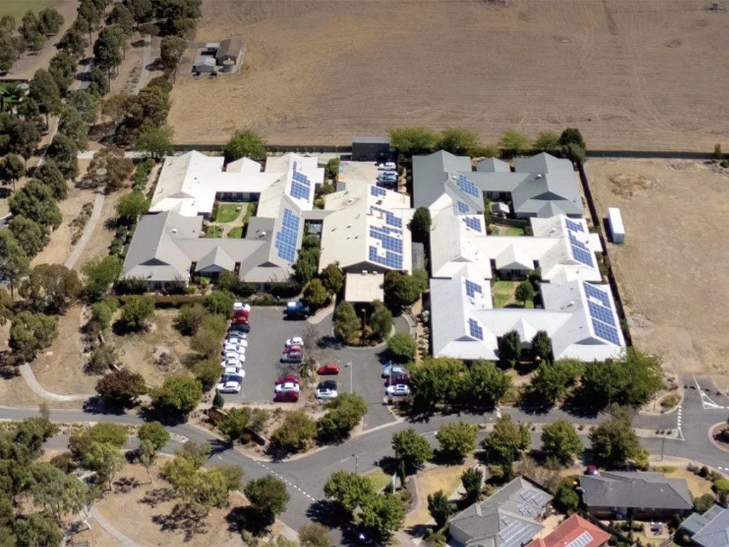 An aerial view of a building complex with solar panels on the rooftops, surrounded by parking lots, trees, and open land.